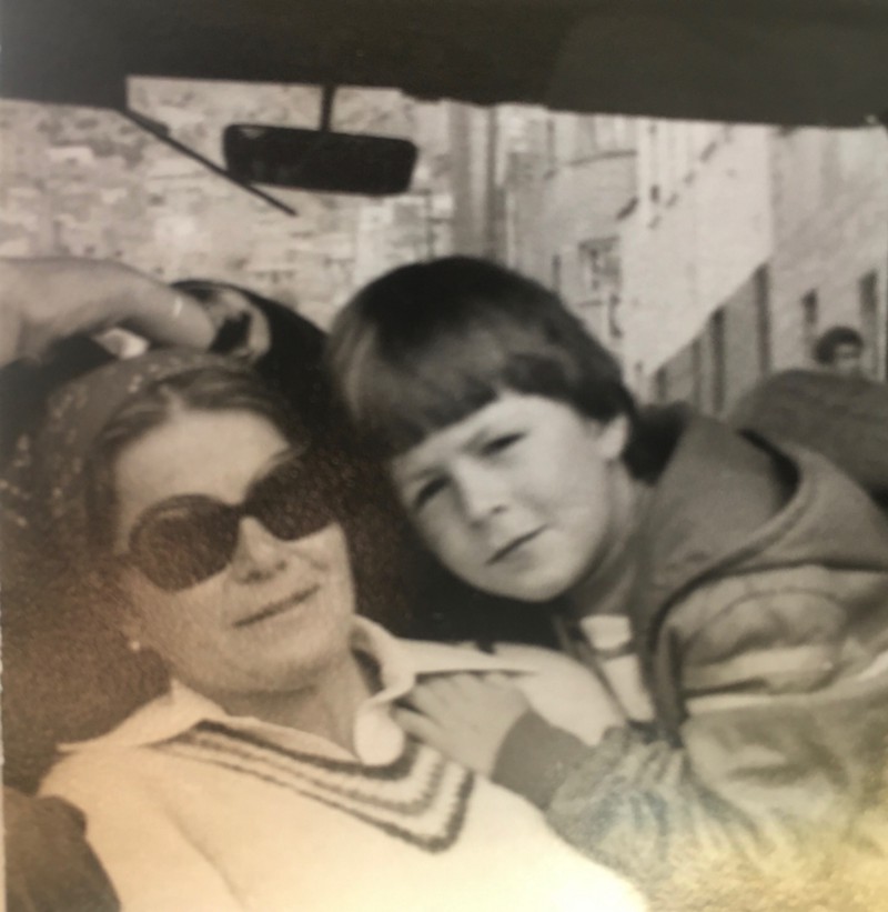 A woman wearing sunglasses sitting in a car, a young child leaning in close, with his hand on her shoulder