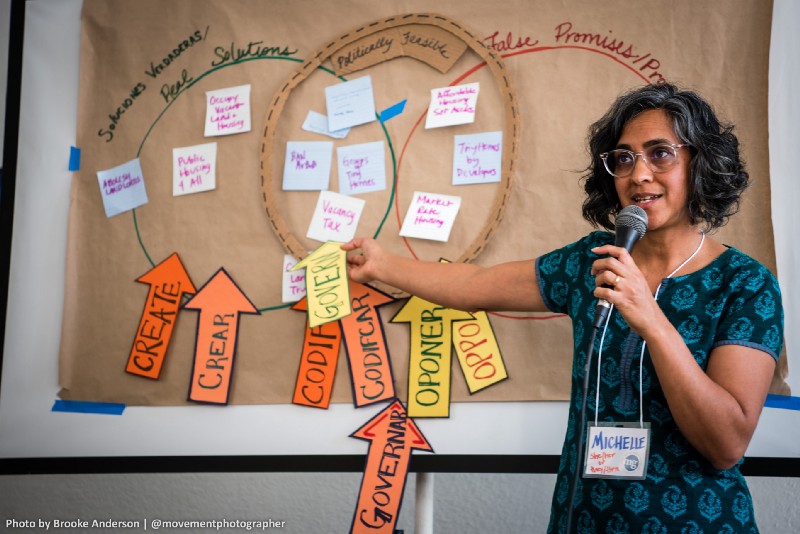 Image of Michelle Mascarenhas, holding a microphone and gesturing towards a butcher paper with a strategy framework drawn on it.
